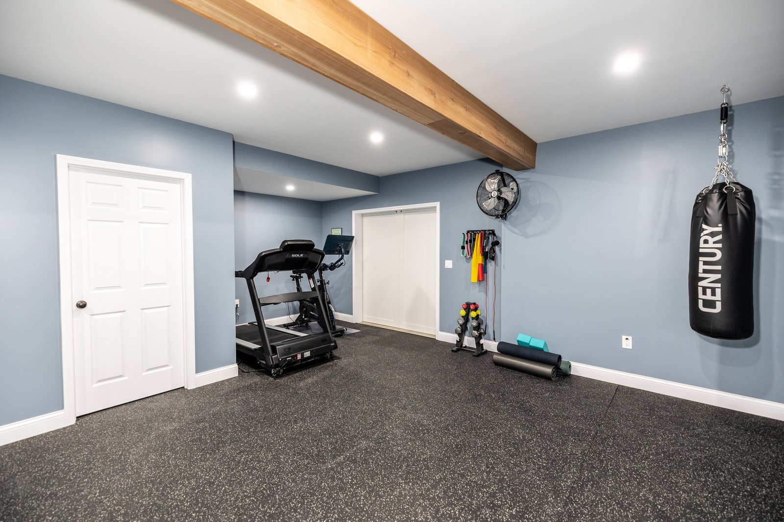 Basement home gym with blue-grey walls, an exposed cedar beam, rubber flooring and a punching bag