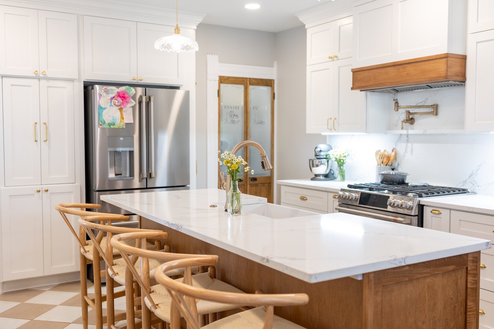 White shaker kitchen with a quartz waterfall island, wood range hood, brass hardware and a checkered tile floor